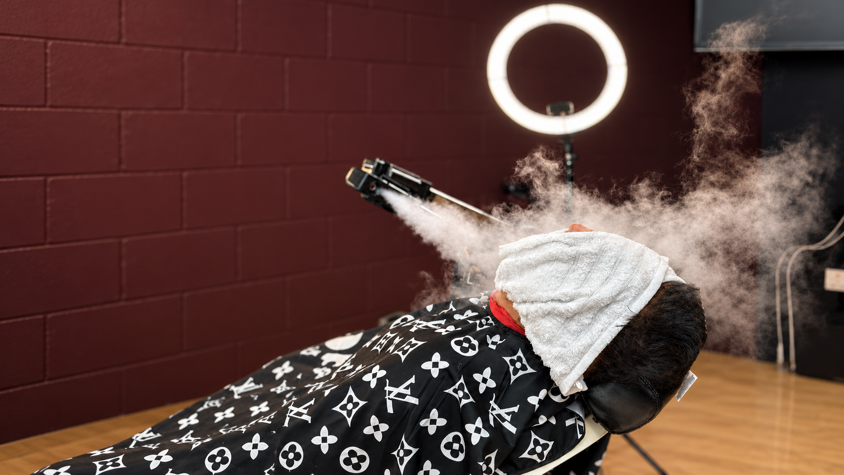 Client reclined for a hot towel steam treatment at God Level Barbershop, steam rising beside the ring light on the burgundy brick wall